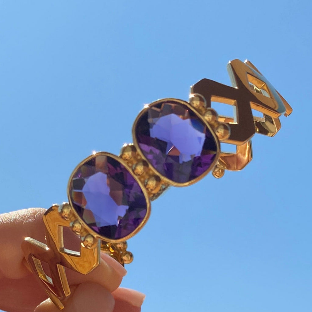 Gold and purple earring held by a hand against a blue sky.